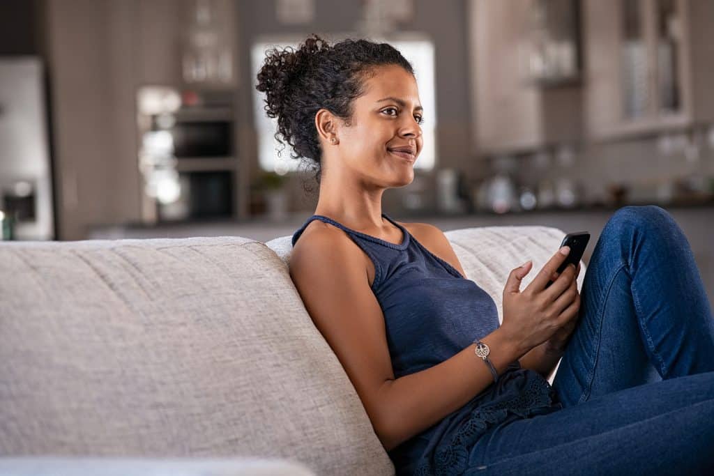 Pensive young woman sitting on couch and holding smartphone whil